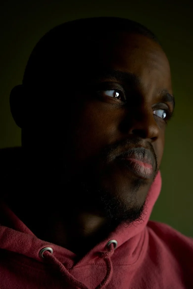 Black man wearing a maroon hoodie looking toward the right of the frame, with a dark, olive green background