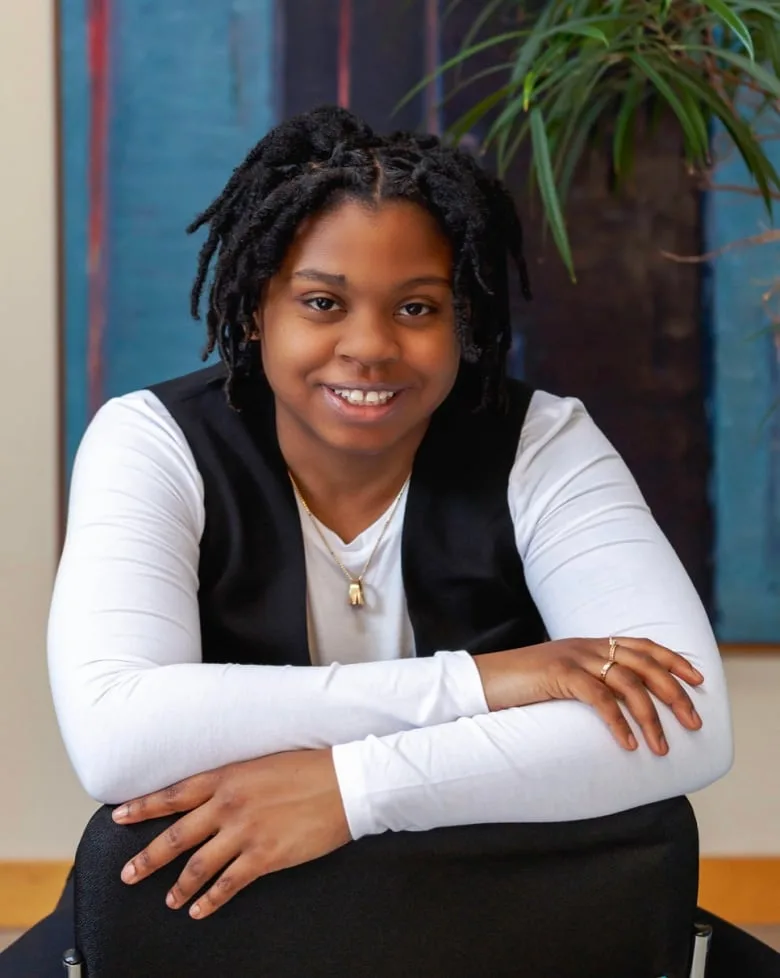 Young Black woman smiles directly into the camera wearing a long sleeve white top with black vest and black pants. Her arms are crossed and there is a small green plant in the background.