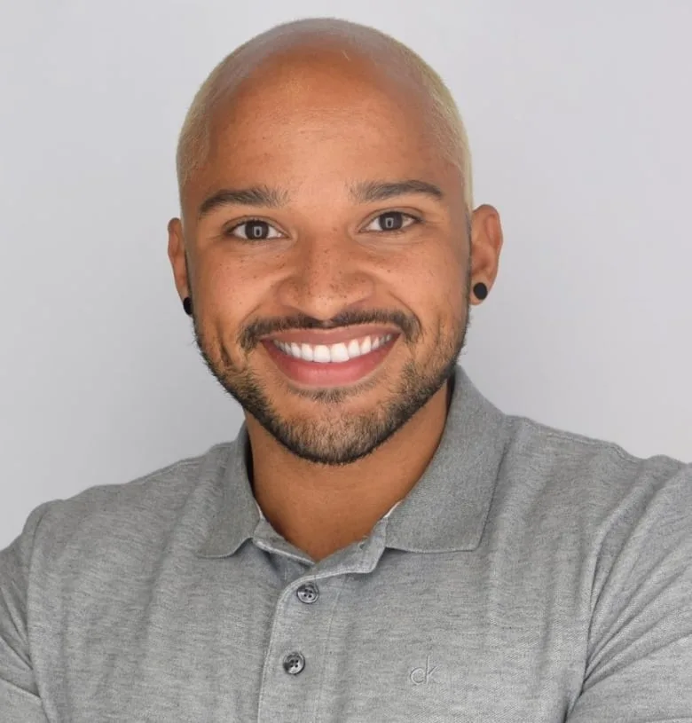 Black man with bald head smiling straight into the camera. He is wearing a light gray Polo shirt and black circle earrings in each ear.