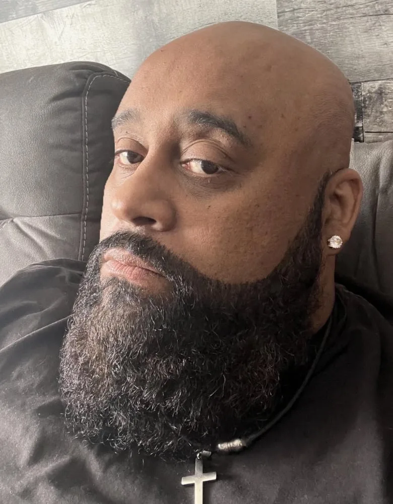 Black man with bald head and dark beard looks into camera. He is wearing a black t-shirt, silver cross necklace and diamond earring in his left ear.