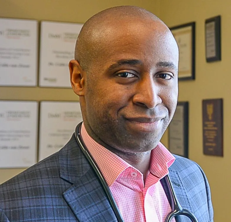 Black man with bald head smiles at camera wearing a blue checkered blazer and pink dress shirt with stethoscope around his neck 