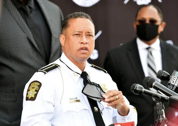 A middle aged man dressed in a white shirt adorned withlaw enforcement patches is speaking to a crowd.