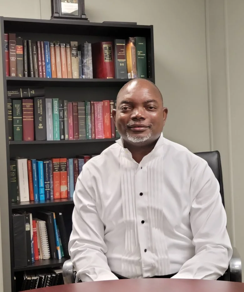 Black man wearing a white dress shirt sits at a table in front of a bookshelf and smiles.