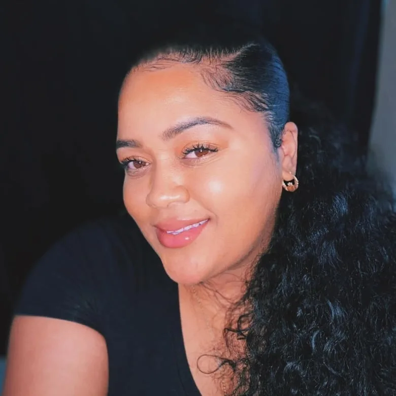 Black woman smiles at camera in front of a dark wall. She is wearing a black t-shirt and has black hair pulled back on a ponytail, and wears small gold hoop earrings.