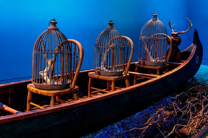 Three wooden chairs sit in a canoe. On each is a bird cage with antlers inside, and behind them sits a shape with antlers on top