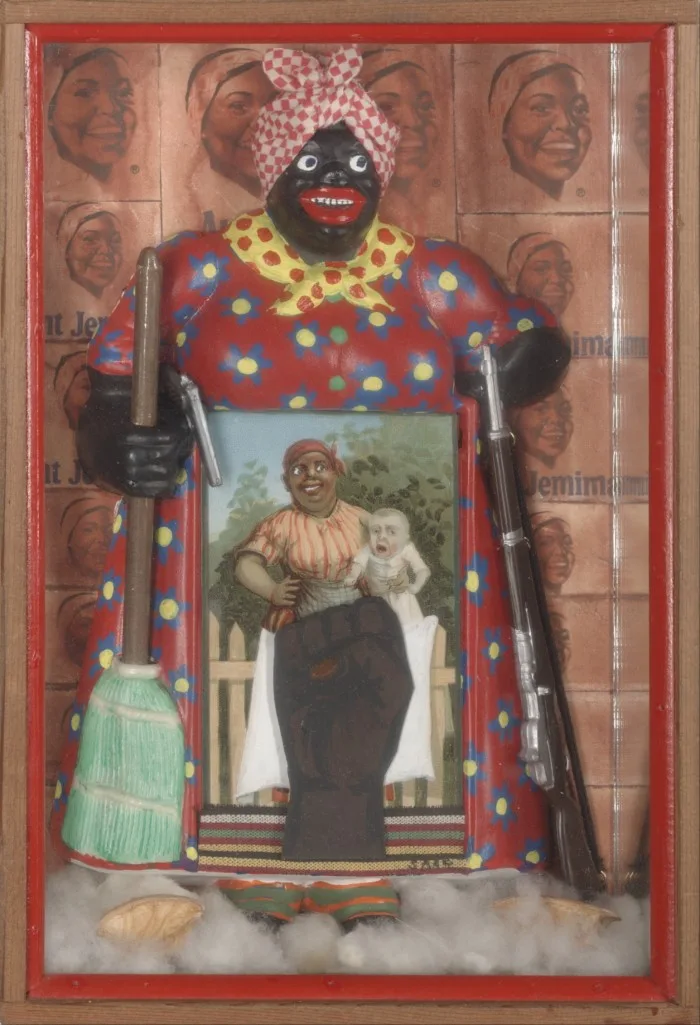 A wooden sculpture of a black woman, depicted as a maid in the pre civil rights South, who holds a broom in one hand and a rifle in the other. In front of her is a picture of another black maid, this time holding a white baby. In front of that is a raised black fist