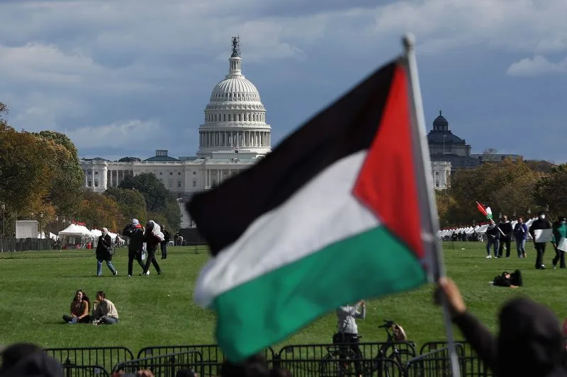 A protester waves a Palestinian flag in front of the U.S. Capitol in Washington, during a demonstration calling for a ceasefire in Gaza. People sit and walk on the grass lawn in front of the protester and barricades.