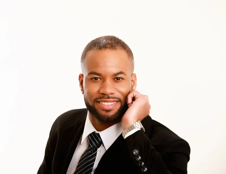 Black man wearing white dress shirt, dark stripe tie and black blazer smiles at camera with his hand to his chin