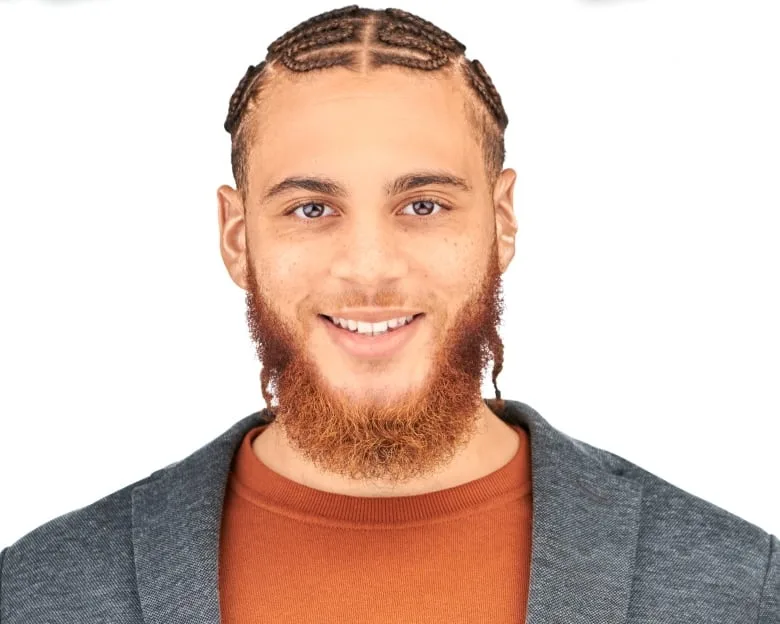 Man with beard, cornrows and goatee wears an orange shirt and gray blazer smiling at camera.