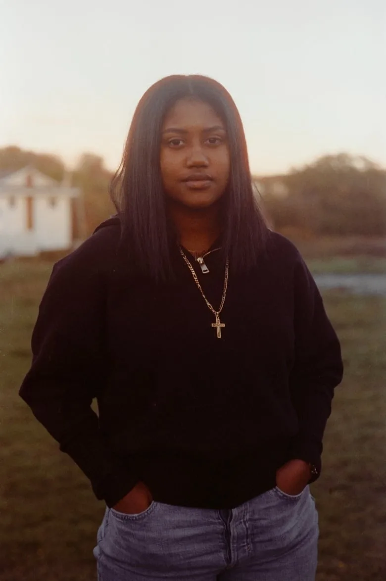 Black woman with long dark hair wearing a black zip-up sweatshirt, gold cross necklace and jeans smiles for the camera