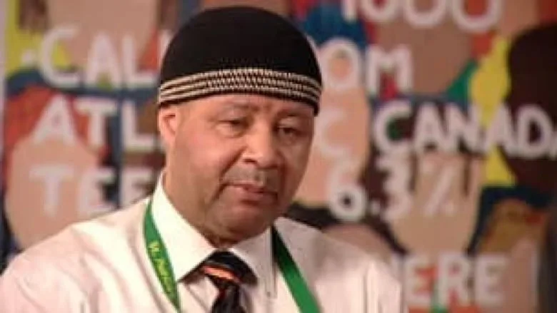 Man wearing black hat with thin white stripes, white dress shirt, black tie, and green lanyard.