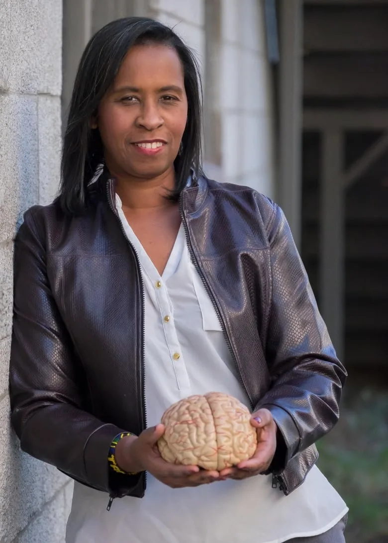 Black woman with long dark hair wearing a white blouse and dark leather blazer holds the model of a human brain in her hands while leaning on a gray wall.