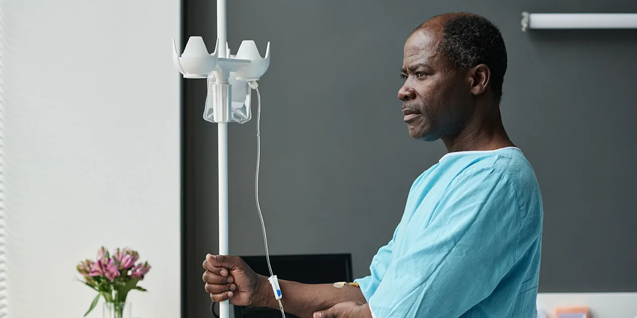 An older Black male patient holding a drip stand and staring into the distance