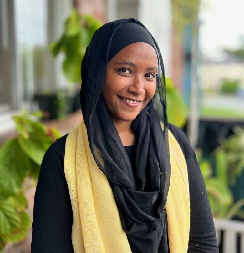 Black woman smiles at camera wearing a black head scarf and robe, with golden scarf over her shoulders.