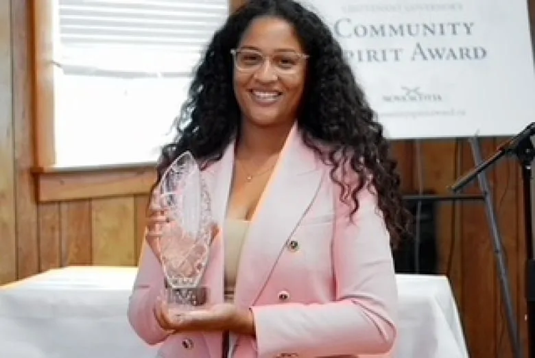 Black woman with long curly hair wearing clear framed glasses, pink blazer and white shirt smiles and holds a clear trophy