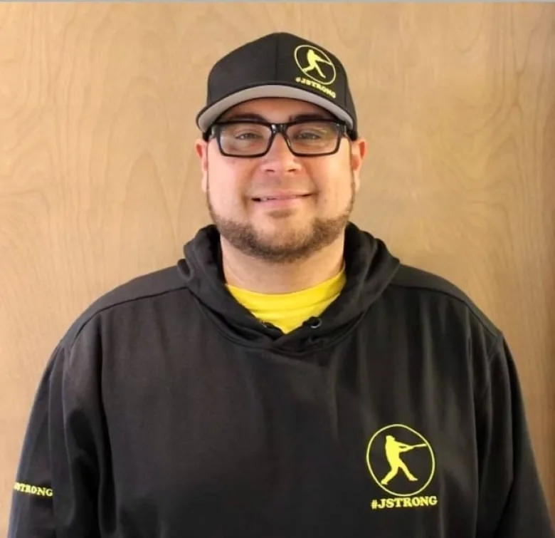 Black man with goatee wearing glasses, black hoodie and black ball cap both with a yellow logo that reads JSTRONG