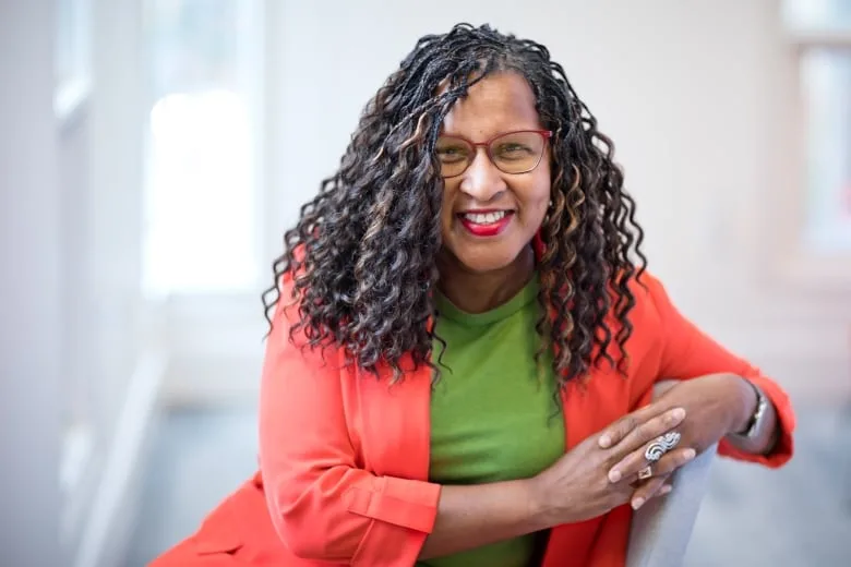 Black woman with long curly hair wears red rimmed glasses, lime green shirt and orange blazer and smiles for the camera