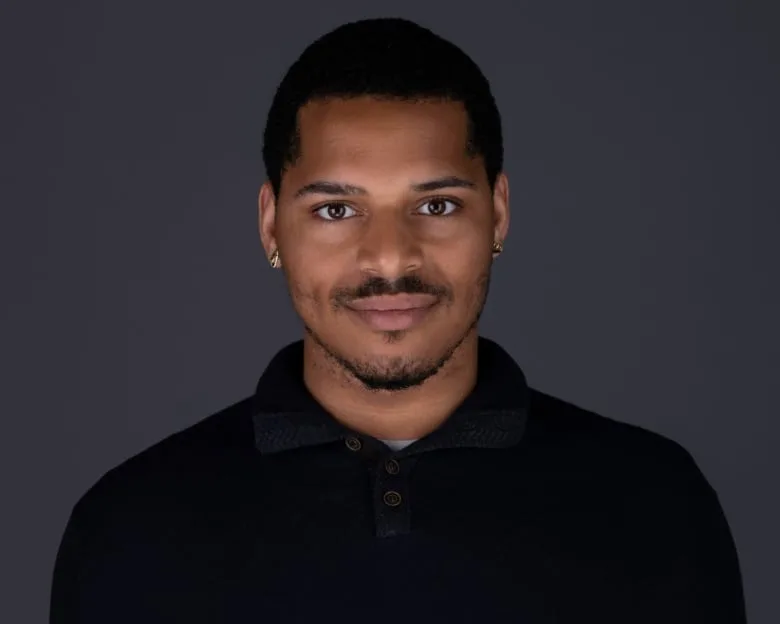 Black man smiling at the camera, wearing a black dress shirt and earrings, in front of a dark background.