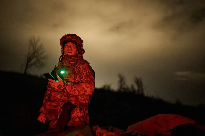 A Ukrainian soldier in helmet and fatigues holds a cell phone and looks up at the night sky as an explosion lights up the horizon behind him.