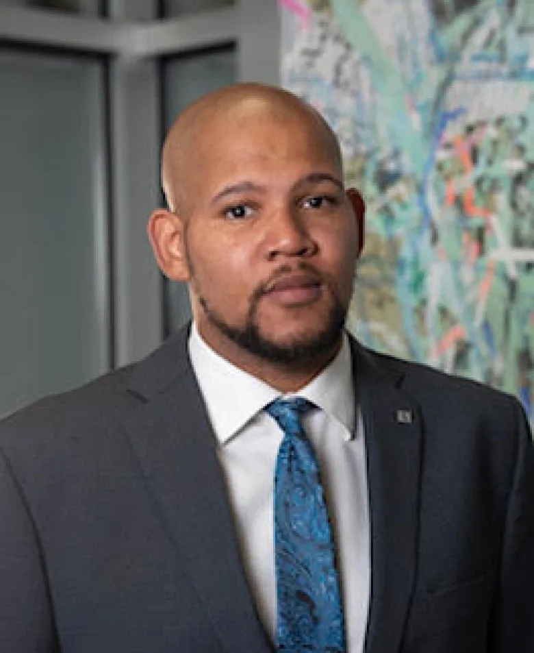 Black man with a goatee smiling at the camera, wearing a white dress shirt, blue tie and blue blazer with a small silver pin on the left lapel. He stands in front of a colourful abstract painting.