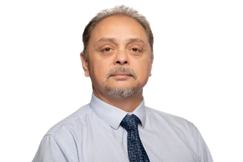 Man with short hair and goatee wearing a white dress shirt with light stripes and blue pattern tie, in front of a white background