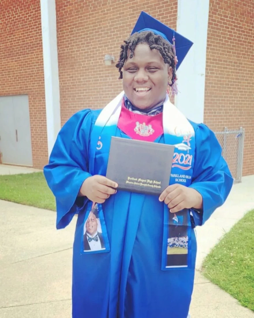 Portrait of Jeremiah Scales, smiling and holding a diploma in a blue graduation gown. 