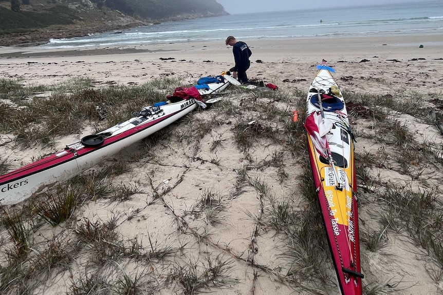 A man in wetsuit moors two kayaks on an island as mist hovers over sea.