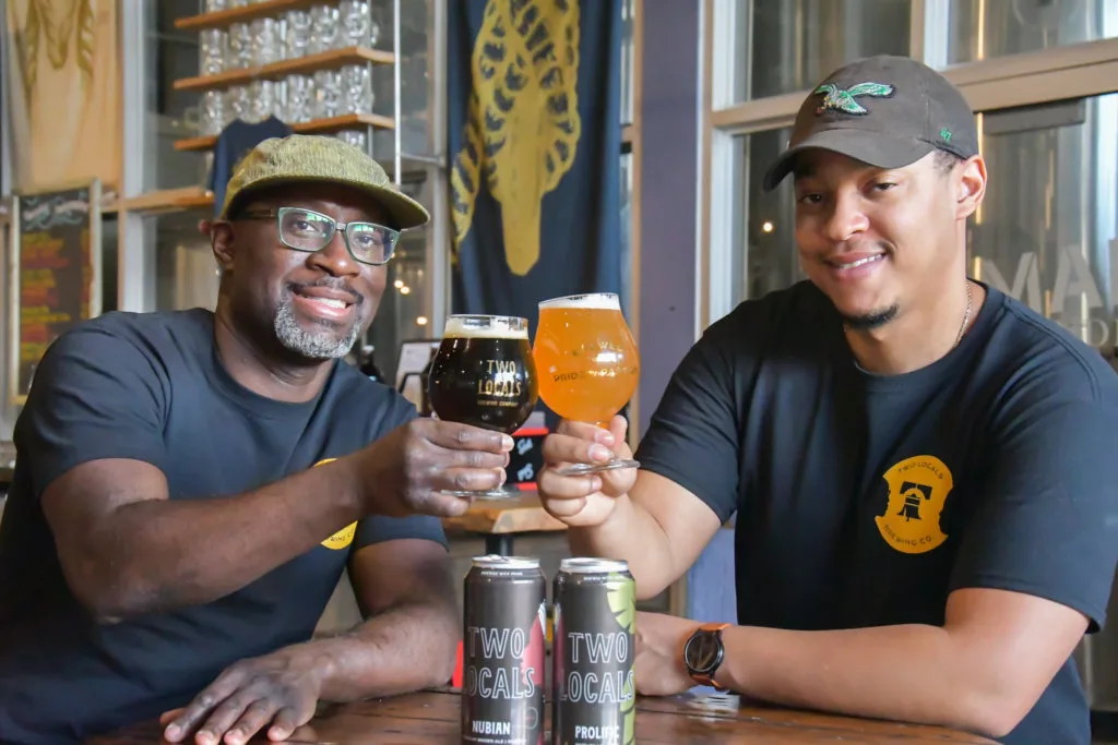 Two men wearing baseball caps smile for the camera while holding beers up to cheers at a bar