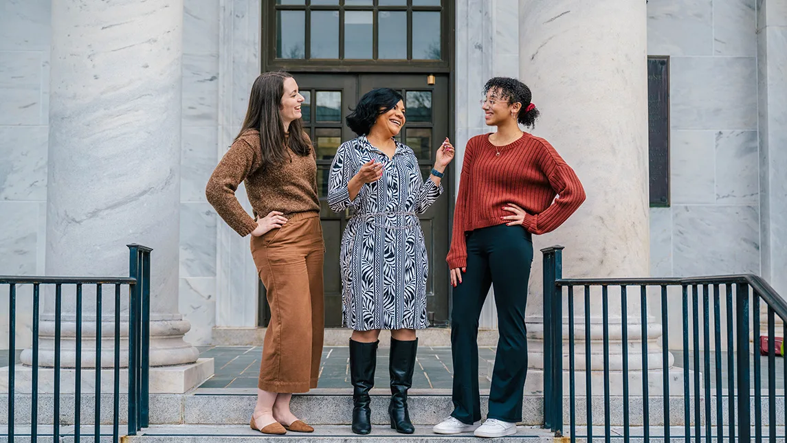 Three women stand outside of a building and talk with one another.