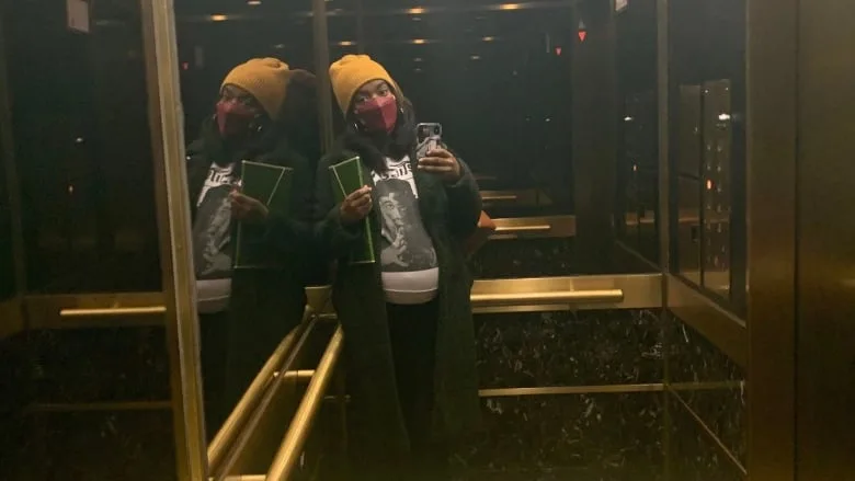 A black woman takes mirror selfie in elevator. She is wearing a cloth face mask and winter clothing. She is visibly pregnant.