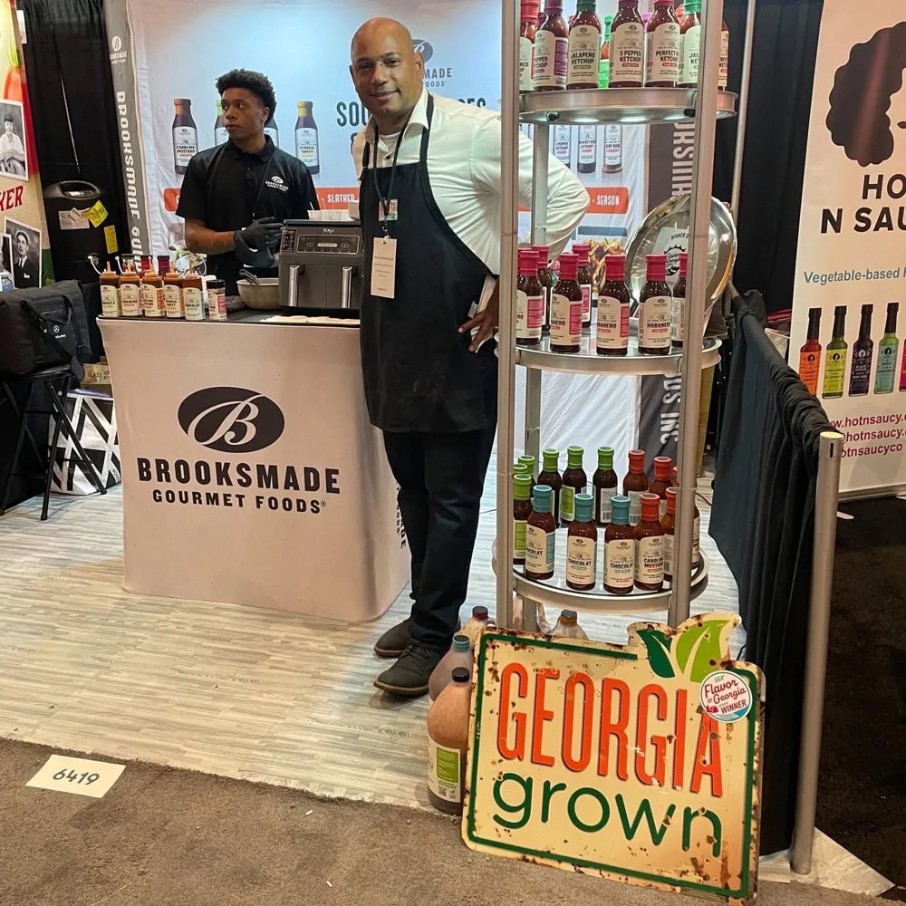 A gentleman in an apron poses in front of a booth with a gentleman standing behind the booth