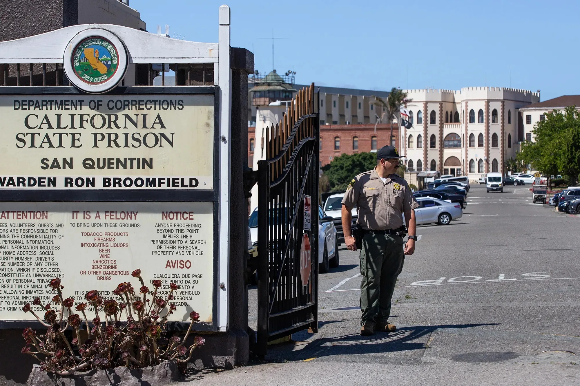 A prison guard in uniform stands in front of a gate with a building in the background.