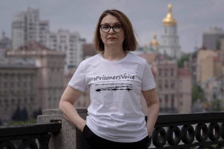 An unsmiling middle-aged woman in a T-shirt saying “Prisoners voice” on a city rooftop 