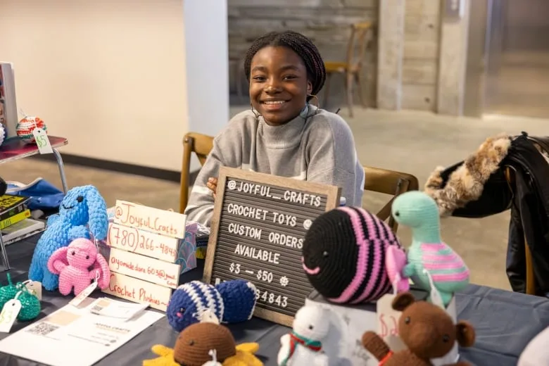 Young woman smiles behind a booth selling crocheted stuffed animals including a black and pink bumble bee and a white unicorn