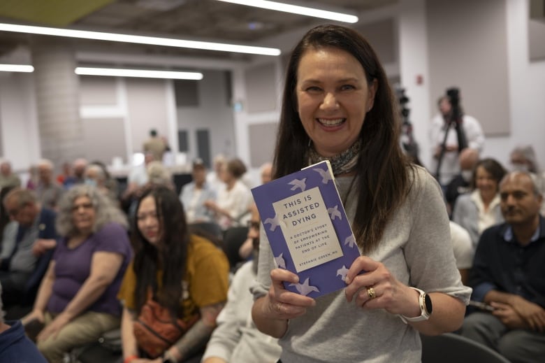 Annette Hamm holds a copy of Dr. Stefanie Green's This is Assisted Dying, in front of a crowd.