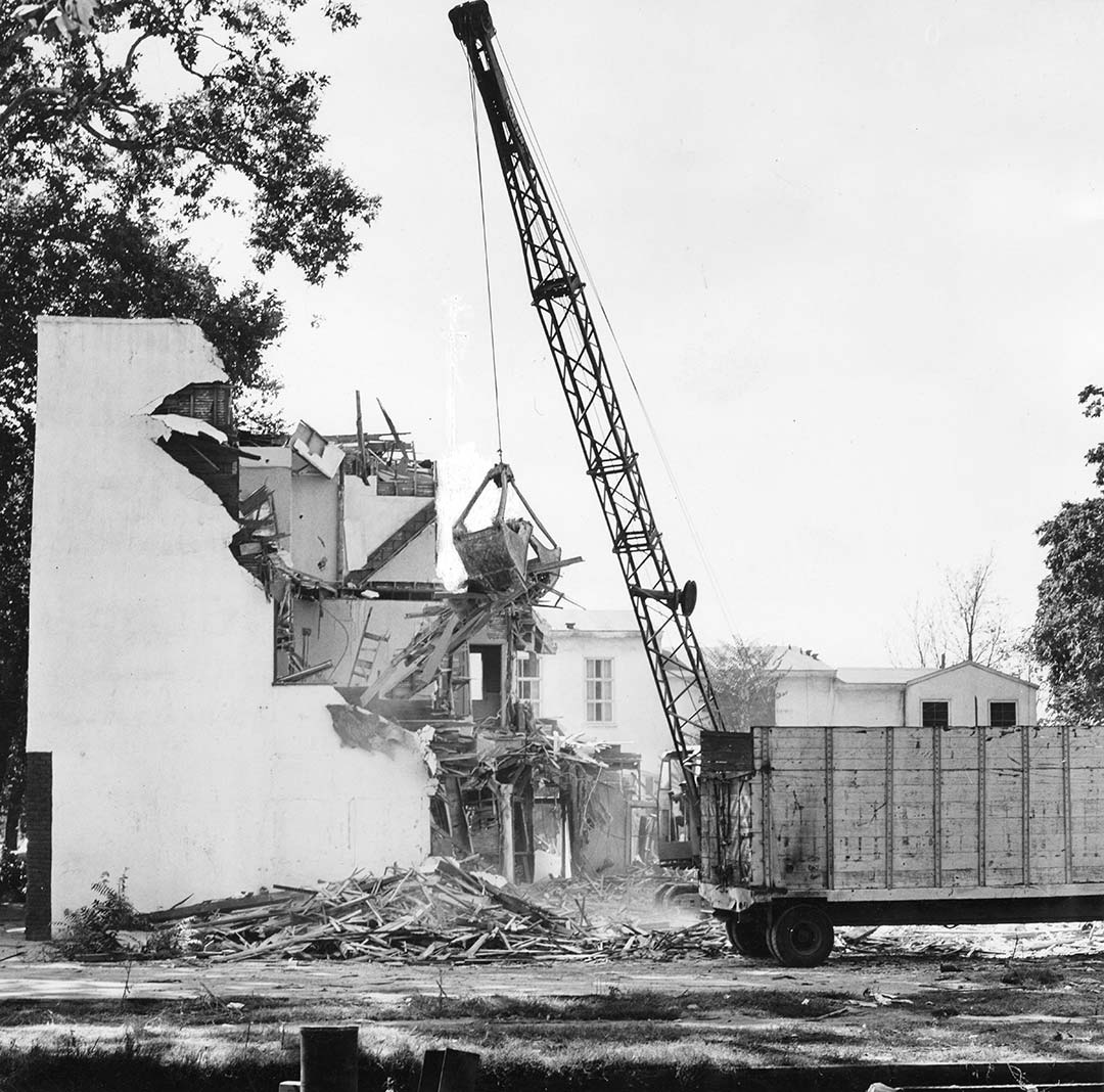 Black and white photo of a building undergoing demolition. A crane with a scoop attachment hauls debris from the partly torn down building to a wooden dumpster.