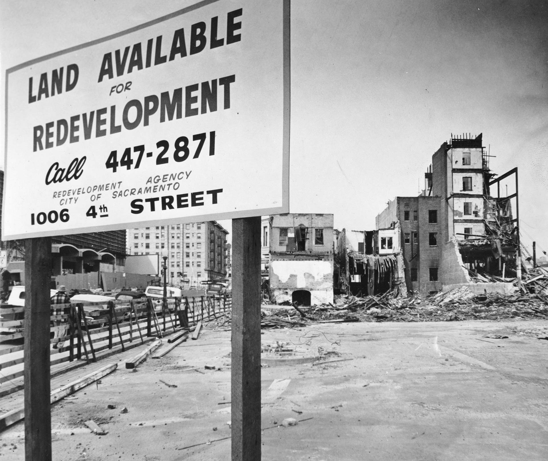 Black and white photo showing a large sign put up in front of a vacant lot. The sign reads 