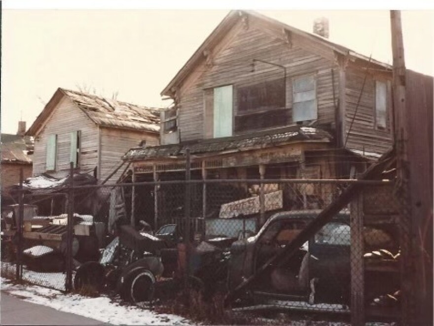 An old photo shows a run down wooden home with broken down cars in the front yard. 