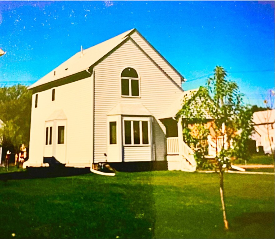 A two story, white wooden home with a large green lawn and a small tree out front. 