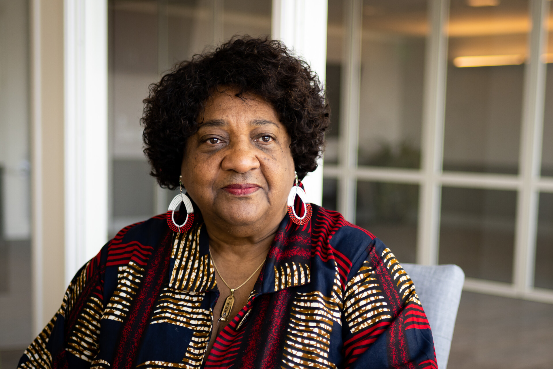 A woman with short, curly brown hair, dangly earrings and a red, blue and cream-patterned blouse sits as she poses for a portrait. A calm look on her face. She wears a simple gold pendant necklace.