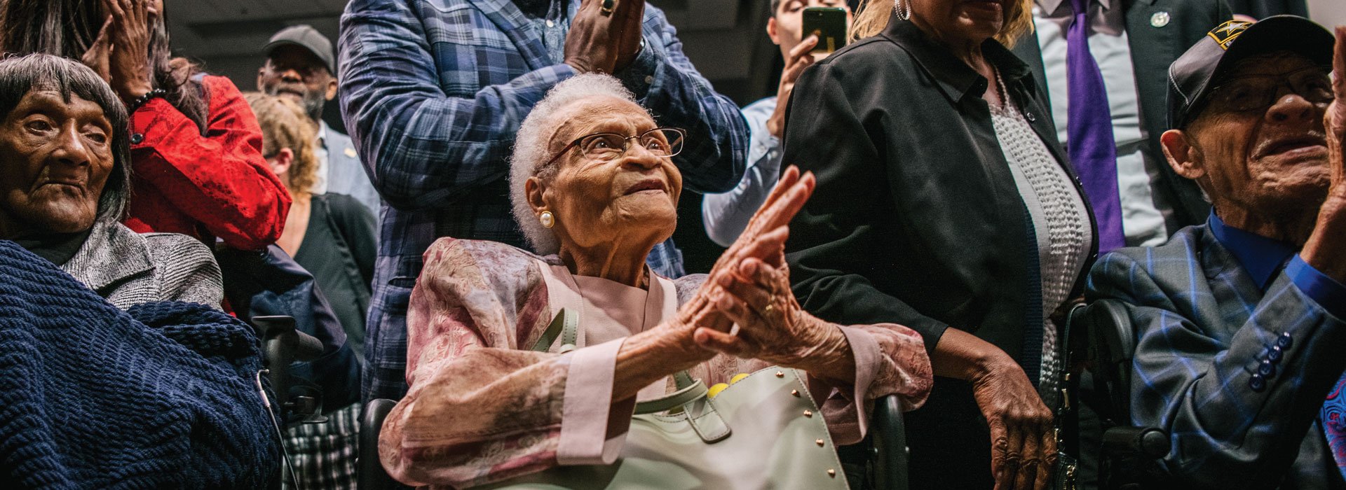 Two elderly Black women and an elderly Black man sit in chairs surrounded by clapping supporters