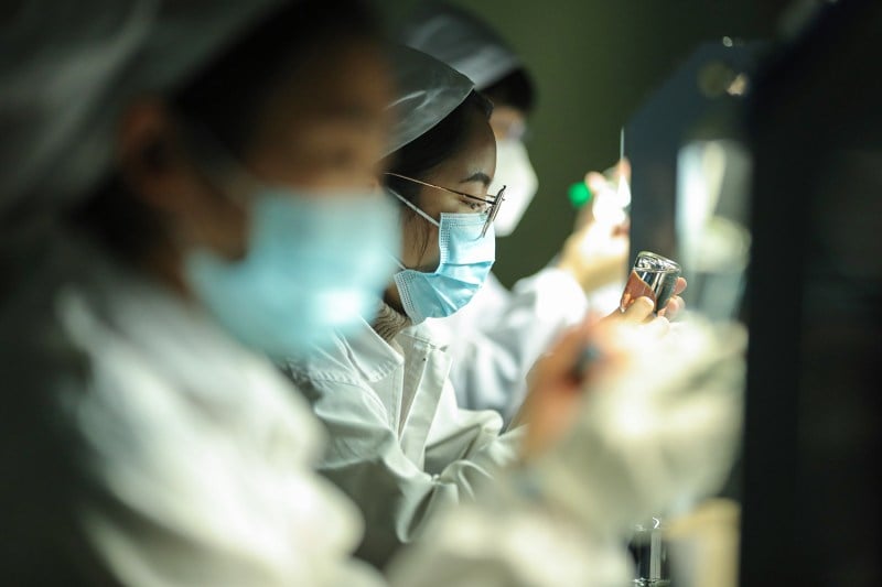 A worker wearing a mask and glasses studies a vial on a pharmaceutical factory assembly line. Other workers are out of focus on either side of her.