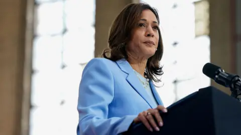 Reuters Kamala Harris wears a blue suit and looks out of frame while standing at a microphone