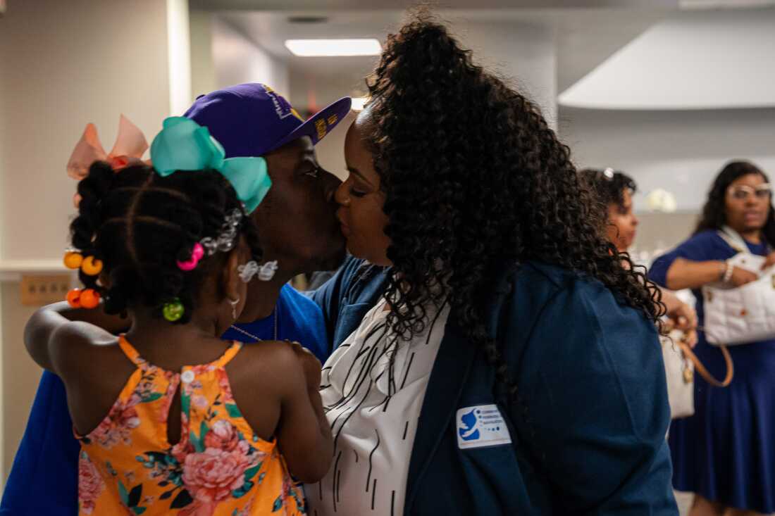 Bristeria Clark kisses her husband while he holds their daughter after the commencement ceremony for Morehouse School of Medicine's first class of rural doulas. Clark is wearing a dark blue blazer and white blouse. Her husband is wearing a blue shirt and blue baseball cap, and their daughter is wearing an orange, flower-patterned dress.