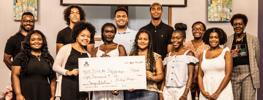 Prince Solace and Willye Bryan flank the 10 scholarship recipients. In the middle, three students hold a large check with $50,000 from the reparations fund going to the scholarships.