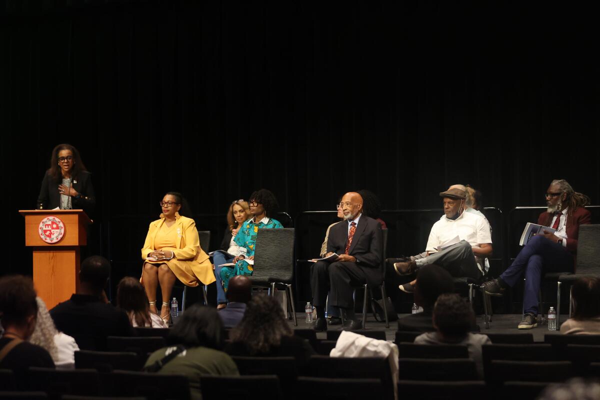 A woman speaks standing at a lectern while others, seated, listen 