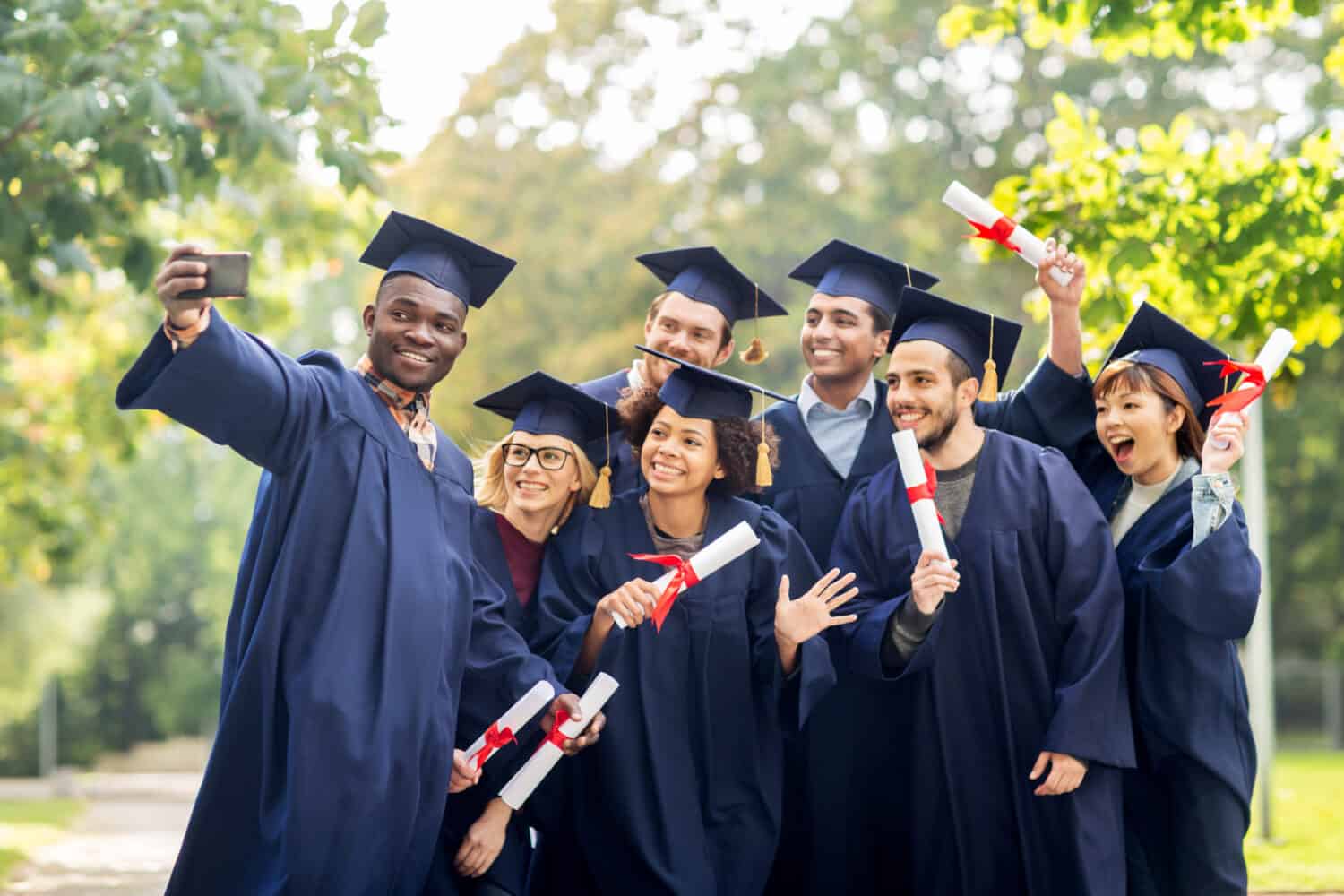 education, graduation, technology and people concept - group of happy international students in mortar boards and bachelor gowns with diplomas taking selfie by smartphone outdoors