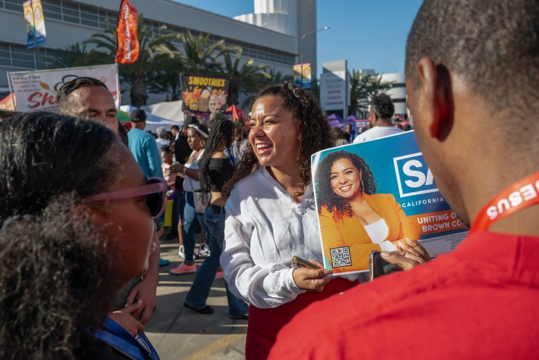 A person standing outdoors in a crowded event holds a poster featuring a political candidate. The candidate, smiling, is pictured wearing an orange blazer with text reading 'Uniting Our Black & Brown Communities.' People are gathered in the background, with food and drink stands, including one selling smoothies. The person holding the poster is engaged in conversation, while others in the foreground face them.