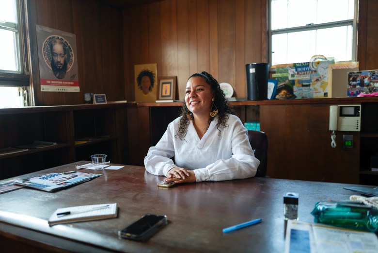 A person sits at a large desk in an office with wood-paneled walls, smiling and wearing a white shirt and distinctive earrings. The desk holds papers, a phone, and a glass of water. Behind them, shelves contain various items, including artwork and a poster on the wall featuring a figure with the text 'The Marathon Continues.' The setting feels warm and personal.