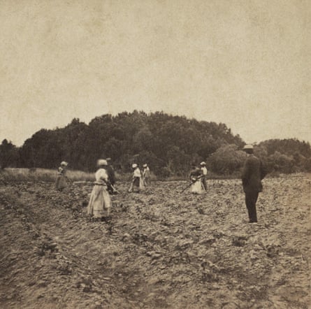 A black and white image showing about six people working on the land. A man in a suit and a hat stands nearby watching them.